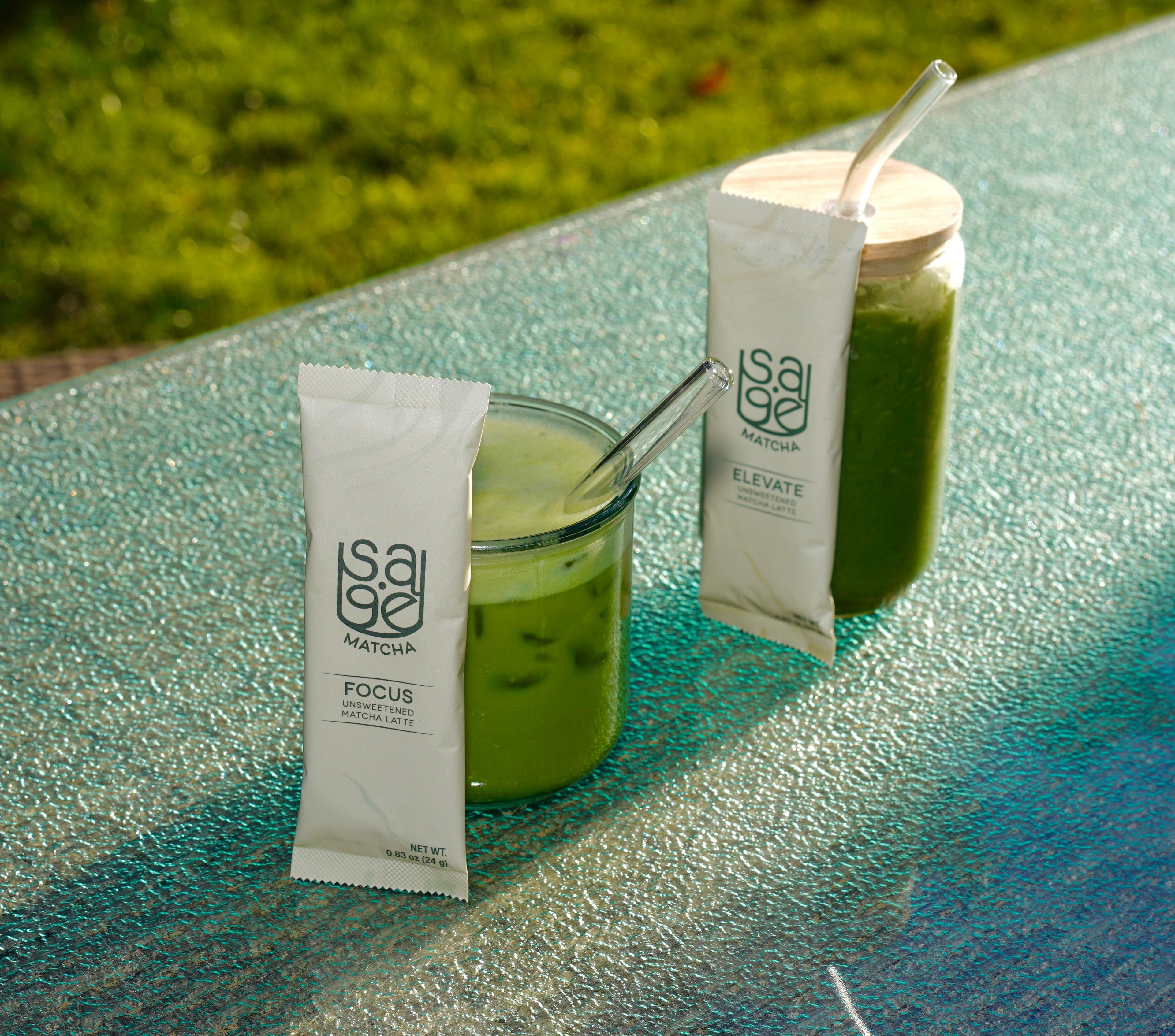 Two glasses of instant matcha lattes, with Sage Matcha sticks in front. Focus Unsweetened and Elevate Unsweetened varieties. Outdoors, nature, glass table top.