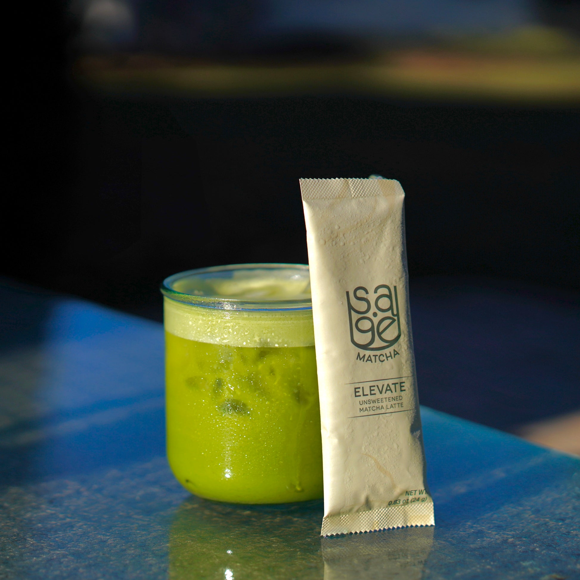 Glass of Sage Matcha latte, single serving stick of Sage Matcha Elevate Unsweetened instant latte leaning against glass, on outdoor glass countertop.