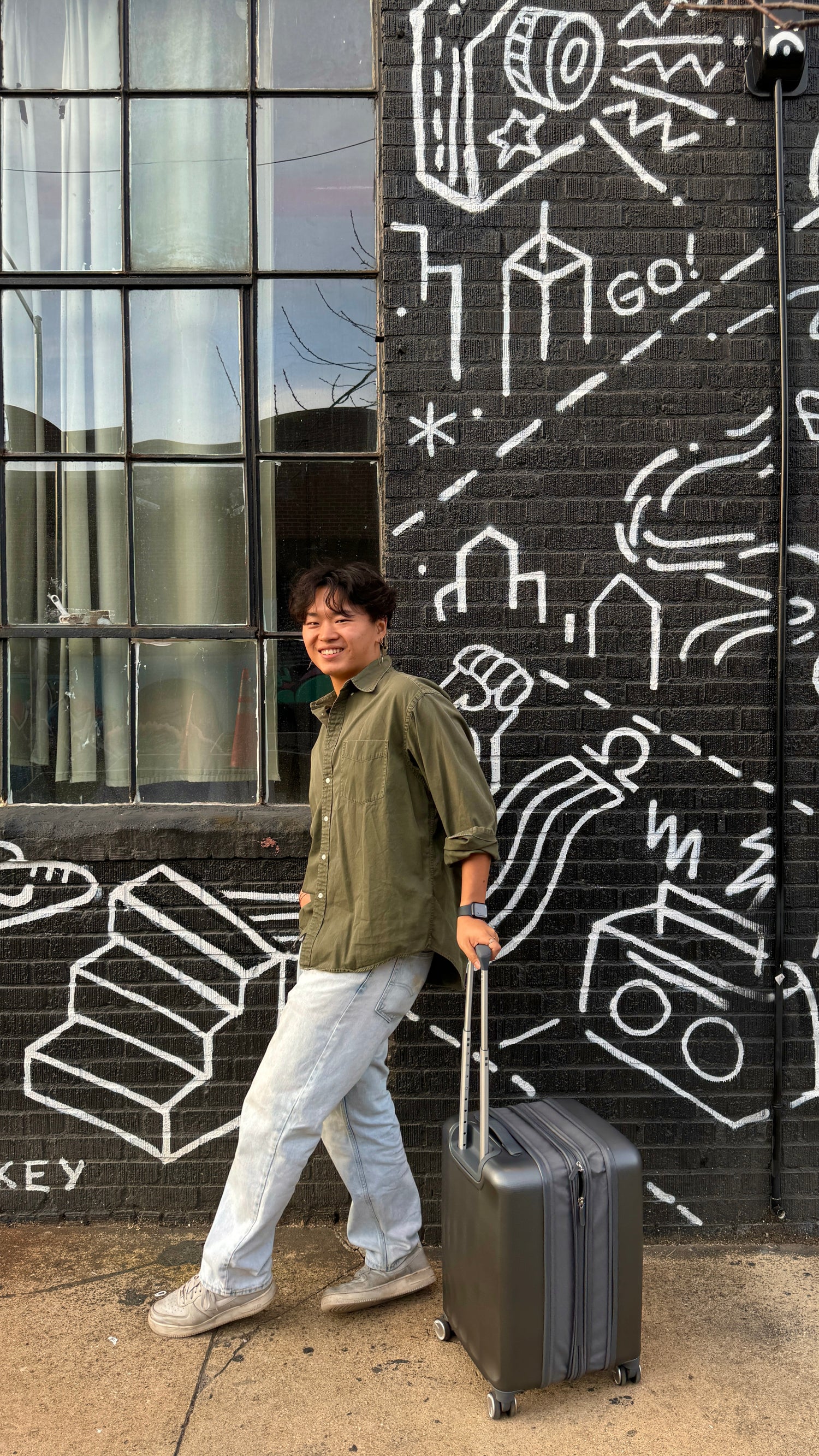 Sage Matcha founder, Larry, holding suitcase and posing in front of graffiti'd black brick wall. 